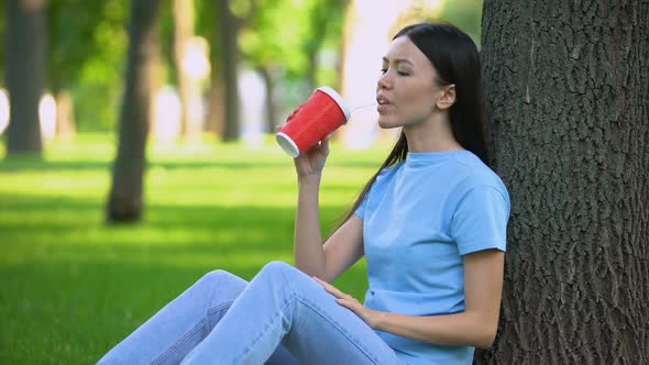 Careless Asian Woman Throwing Away Empty Paper Glass Siting Under Tree in Park alt