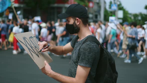 Masked riot activist man drawing poster sign, poster banner. Rebel on rally. alt
