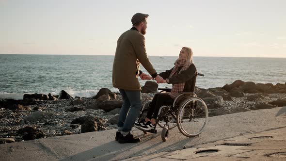 Joyful Man Is Holding Hands of His Disabled Girlfriend on Embankment of Sea