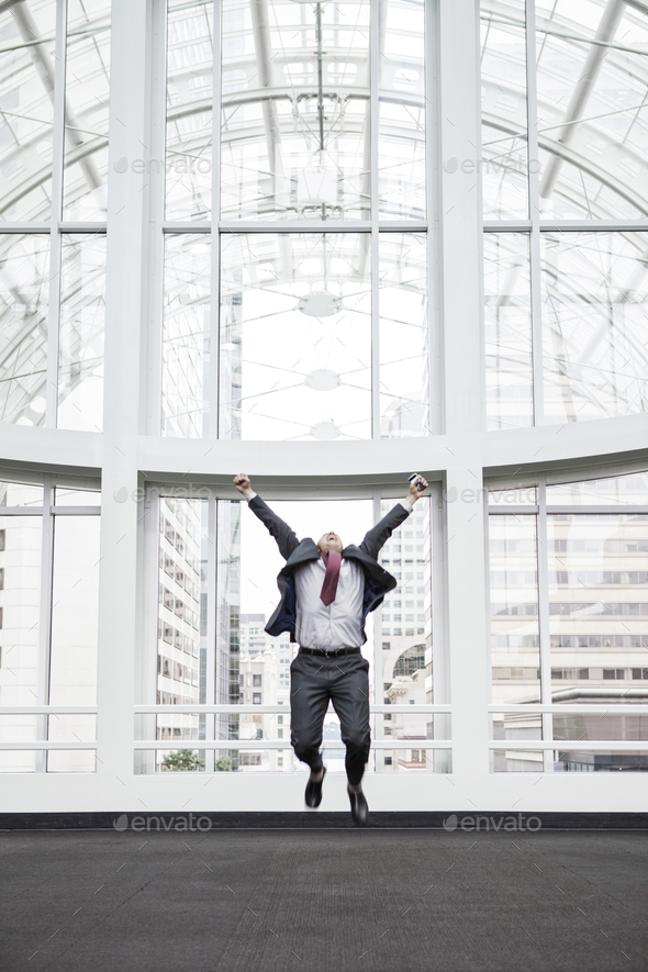 A businessman jumping for joy in front of a large window in a convention center lobby.