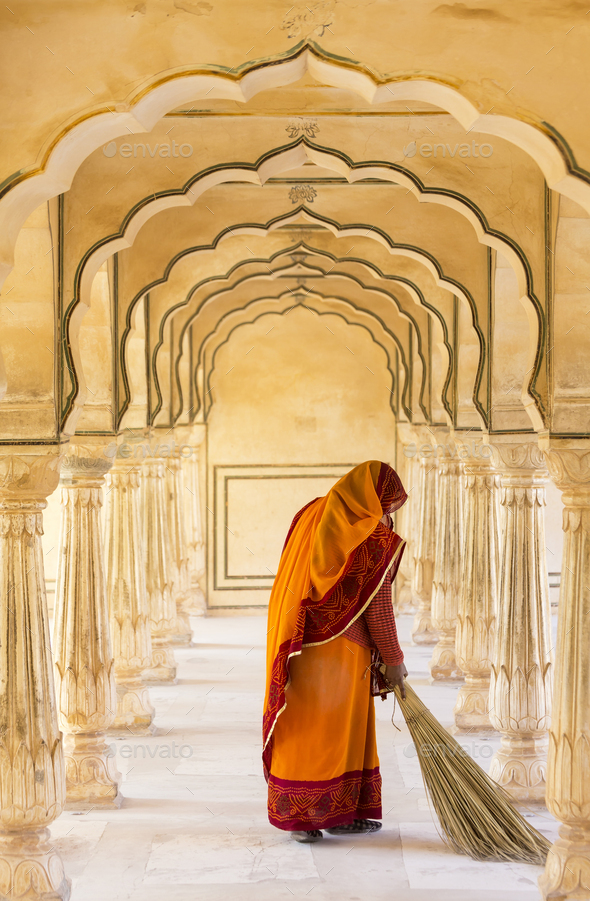 Rear view of woman wearing orange sari using broom to sweep floor of a colonnade.