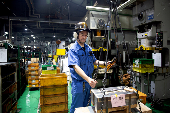 Japanese man wearing safety helmet, ear protectors and blue overall standing in factory, working.