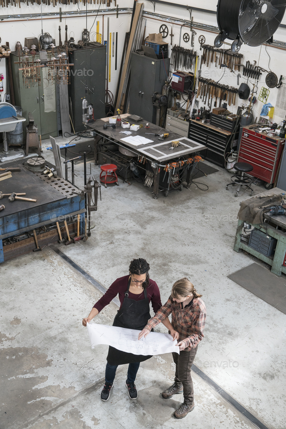 High angle view of two women standing in metal workshop, holding technical blueprint.