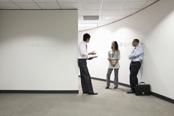 A mixed race group of three businesspeople standing and talking in an office hallway.