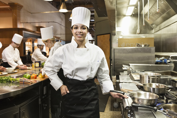 A portrait of a young black female chef in a commercial kitchen with her crew working behing her.