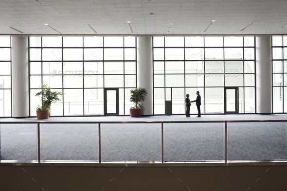 Two businessmen silhouetted and shaking hands in front of a large convention center window.