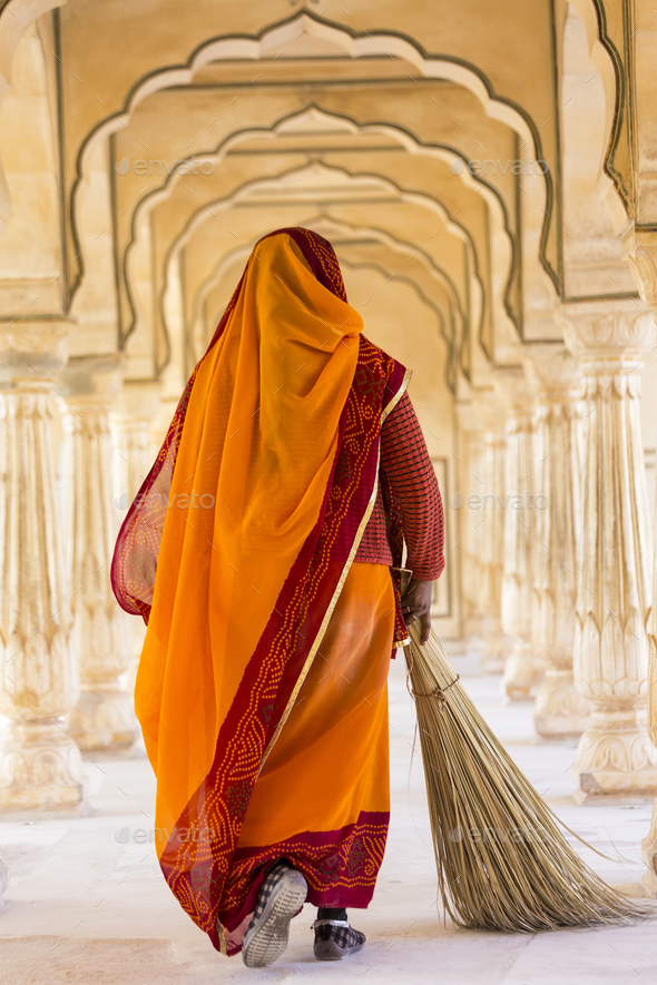 Rear view of woman wearing orange sari using broom to sweep floor of a colonnade.