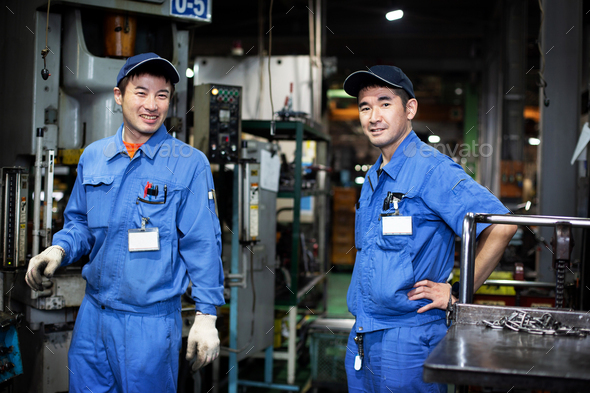 Two Japanese men wearing baseball caps and blue overalls standing in factory, smiling at camera.