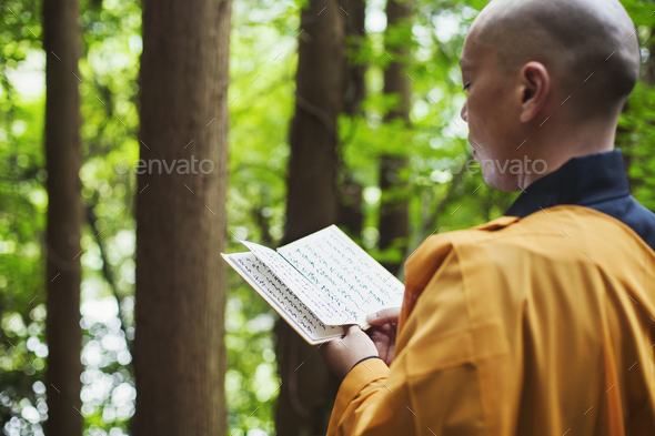 Side view of Buddhist monk in robes standing outdoors, holding prayer text.