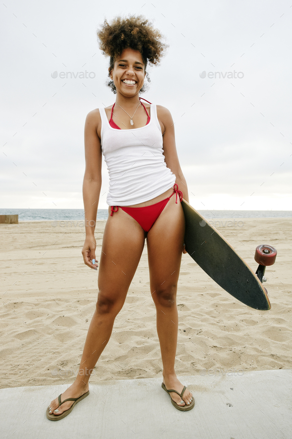 Young woman wearing white vest and red bikini standing on sandy beach, holding skateboard.