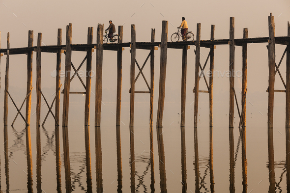 Two people cycling across tall teak bridge over a lake. Stock Photo by  Mint_Images