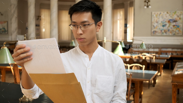 Asian male student intently opening envelope with exam results in university library