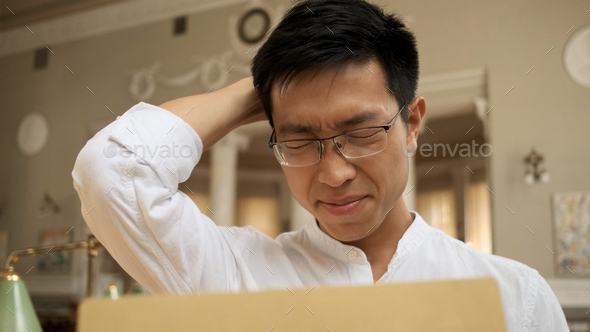 Upset asian male student opening envelope with exam results in library and sadly holding head