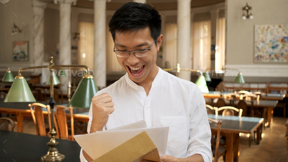 Portrait of asian student joyfully opening envelope with exam results in university library