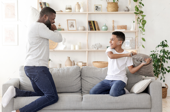 Happy Time Together. Black Dad Pillow Fighting With Preteen Son At Home