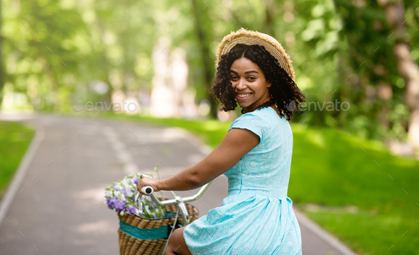 Happy black girl in summer dress riding bike at beautiful green park, copy space