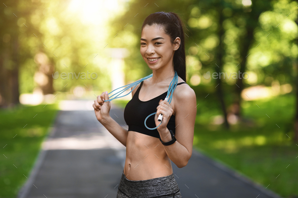 Fit smiling asian woman posing with skipping jump rope outdoors