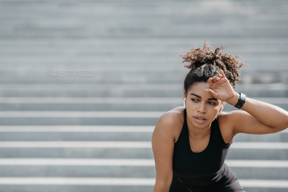 Tired girl in sportswear and smart watches wipes sweat from forehead on stairs background