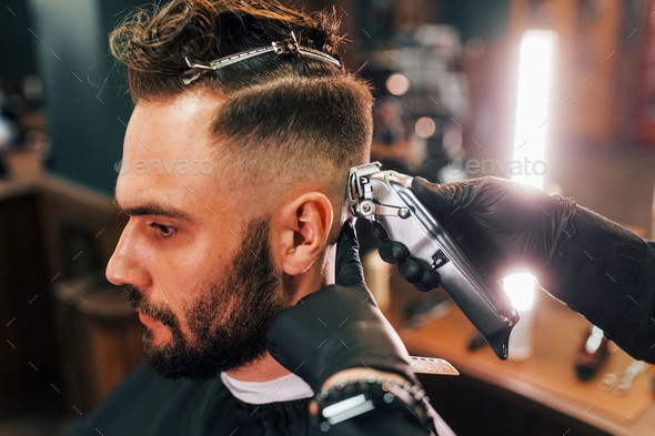 Close up view of young bearded man that sitting and getting haircut in barber shop