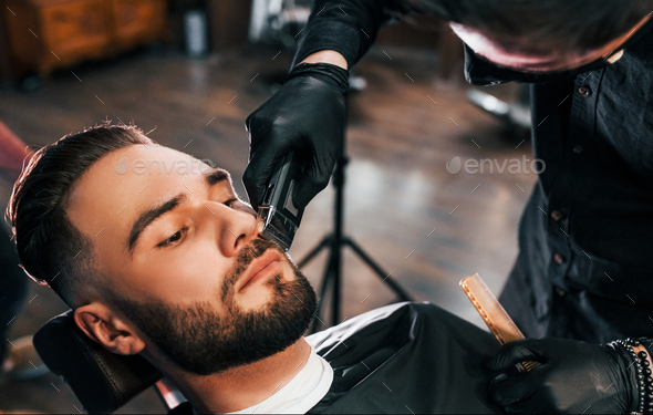 Young man with stylish hairstyle sitting and getting his beard shaved in barber shop
