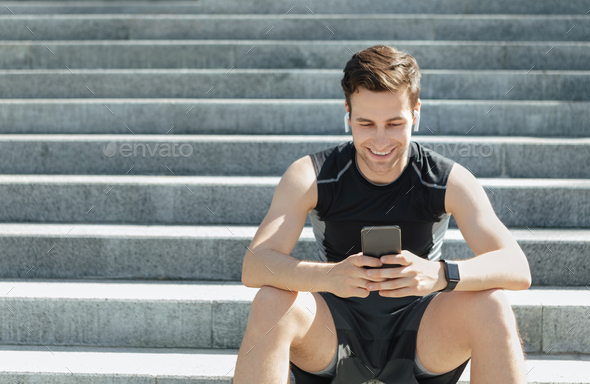 Smiling man with wireless headphones and fitness tracker, sits on city stairs and typing on