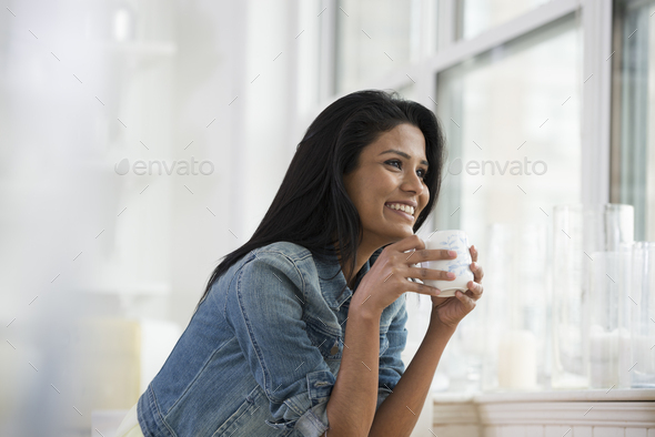 A room interior in the city. A young woman wearing a denim shirt, holding a china cup.