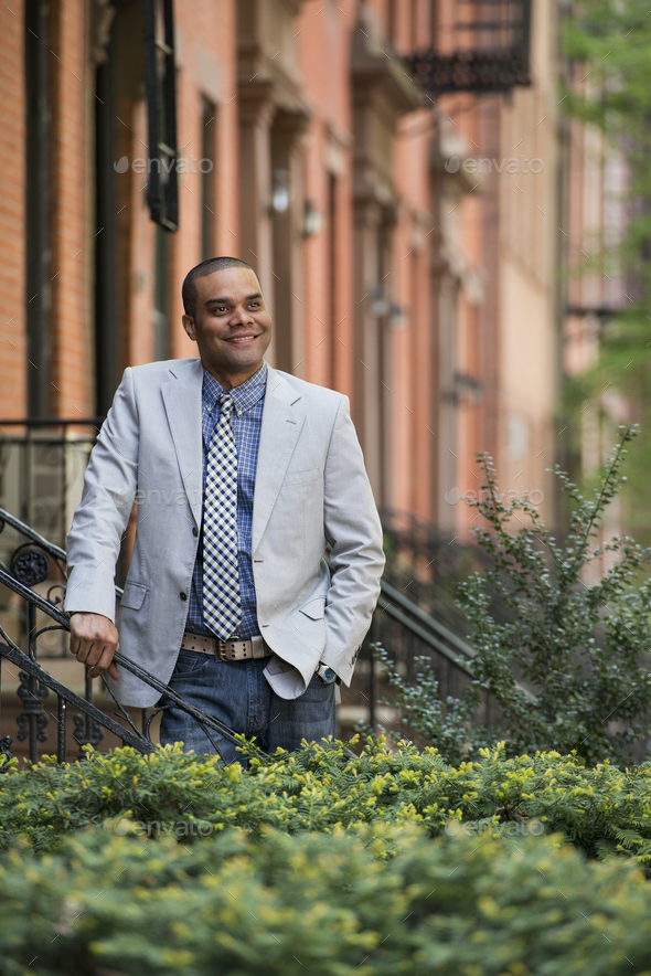A man in a jacket and tie on a city street outside a terrace of brownstone houses.