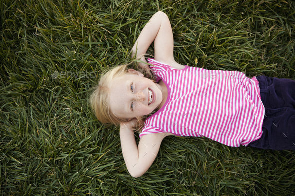 A child, a young girl lying on her back on the grass, with her hands behind her head, smiling and looking at the camera. View from above.