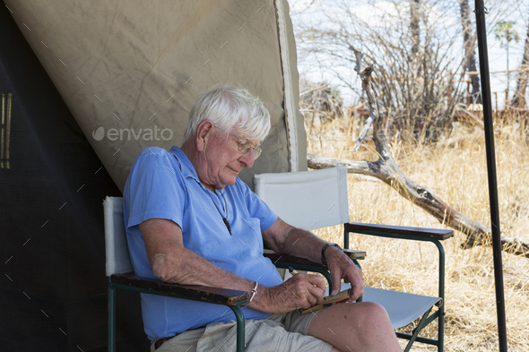 Senior man writing a journal seated outside a tent in a wildlife safari camp.