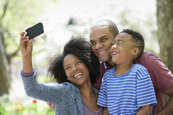 A family in the park on a sunny day. Two adults and a young boy taking photographs with a smart phone.