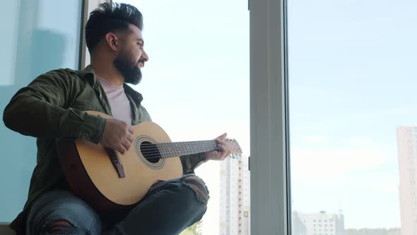 Slow Motion of Young Middle Eastern Man Playing the Guitar Singing Song Sitting on Window Sill at alt