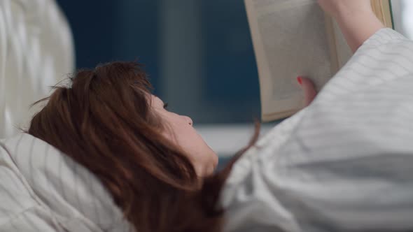 Brunette Woman Lies on the Bed and Reads a Book in the Evening