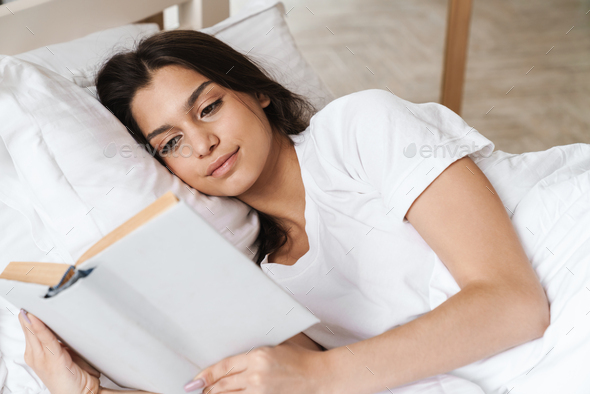 Photo of focused beautiful woman reading book while lying on bed