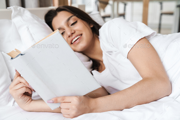 Photo of focused smiling beautiful woman reading book while lying on bed