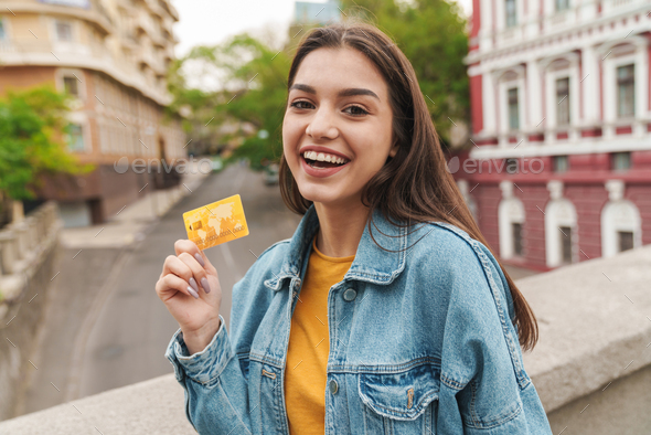 Image of young woman smiling while showing credit card while walking