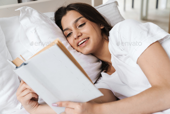 Photo of focused smiling beautiful woman reading book while lying on bed