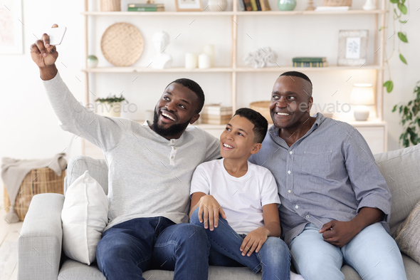 Smiling black father, son and grandfather sitting on couch and taking selfie