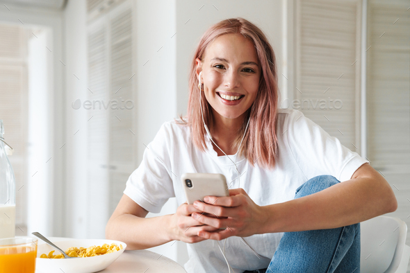 Photo of woman using cellphone and earphones while having breakfast