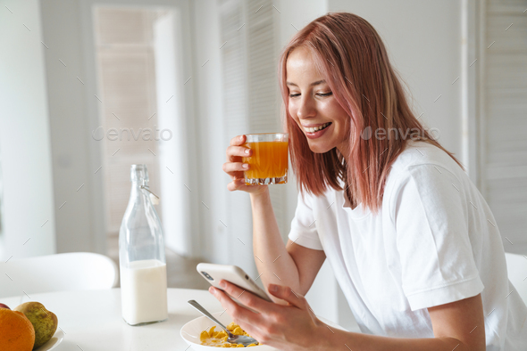 Photo of young joyful woman using cellphone while having breakfast