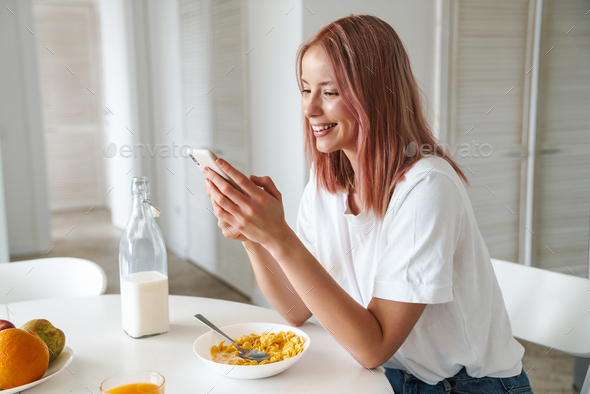 Photo of young cheerful woman using cellphone while having breakfast
