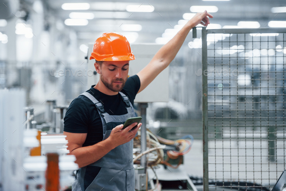 Smartphone in hand. Industrial worker indoors in factory. Young technician with orange hard hat