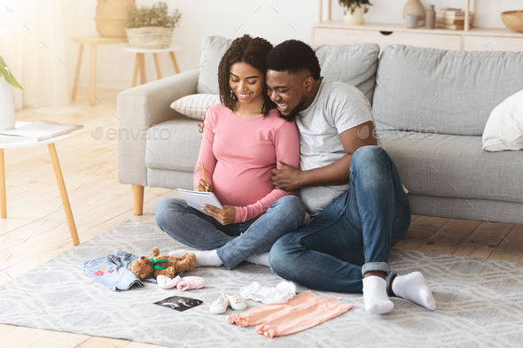 Cheerful pregnant black family preparing baby clothes and making list