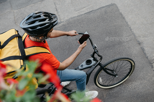 Bicycle courier in protective helmet with delivery backpack sits on bicycle and looks at phone and