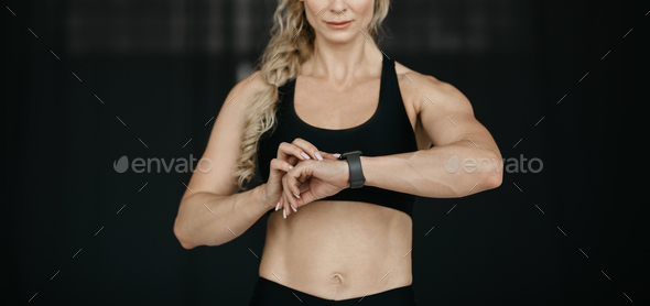 Training result. Muscular adult woman in sport bra, looks at fitness tracker on arm on dark