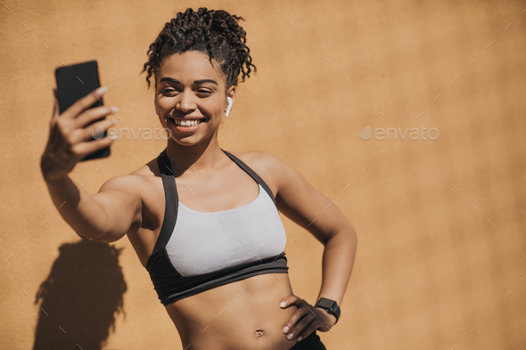 African american young woman in sports bra with wireless headphones and fitness tracker making