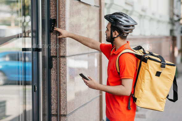 Modern food delivery. Young man with beard, with backpack and smartphone, ringing the bell