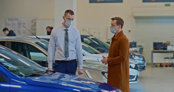 Car Dealership Consultant Wearing a Medical Mask Shows a New Auto in a Luxury Car Dealership