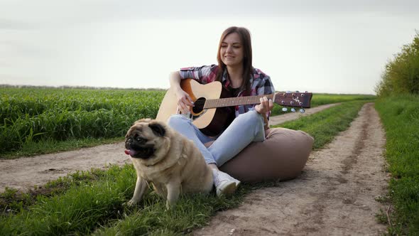 Woman Singing and Playing Guitar with Pug Dog Sitting on Bag Chair in Green Field alt