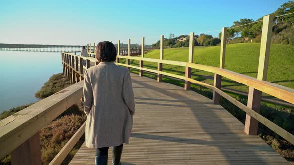 Two Females Walking on a Boardwalk Near a River Shore with People Ahead alt