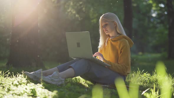 A Young Pretty Woman in a Bright Yellow Sweater Sits on the Green Grass and is Typing on the Laptop